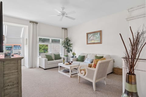 The living room offers a relaxed, coastal-cottage feel with soft neutral tones, a mix of cozy seating, and plenty of natural light pouring in through tall windows.