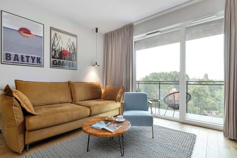 A modern living room features a tan velvet sofa on a patterned rug next to a light blue armchair. Large sliding glass doors lead out onto a sunny balcony.