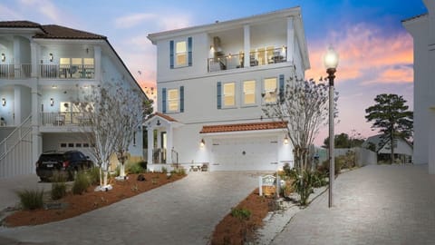 Elegant three-story coastal home with tile roof and spacious balconies, set in a tranquil neighborhood beneath a stunning sunset sky.