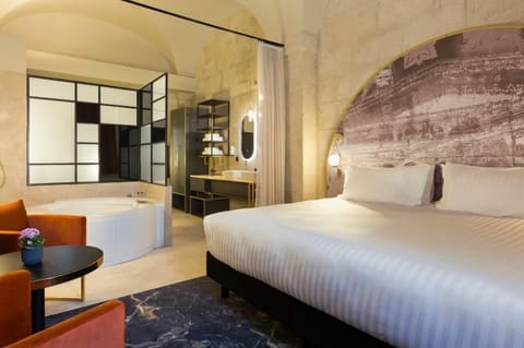 The deluxe room featuring a king-size bed, a dramatic arched fresco headboard wall, and an open-plan bathroom behind elegant black glass partitions.