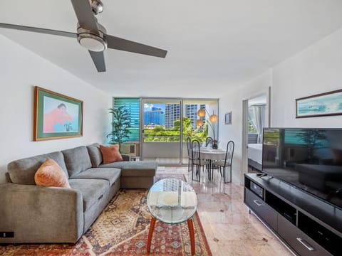 Living Room - Welcome to Harbor View Plaza #409, a bright two-bedroom Waikiki monthly rental with a comfortable sectional, 65-inch Smart TV, dining area, and private lanai/balcony framed by palms and city views.