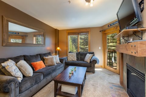 A living room with two gray couches, a wooden coffee table, a TV mounted above a fireplace, and large windows with a forest view. Pillows are arranged on the couches, and a plant decorates the table.
