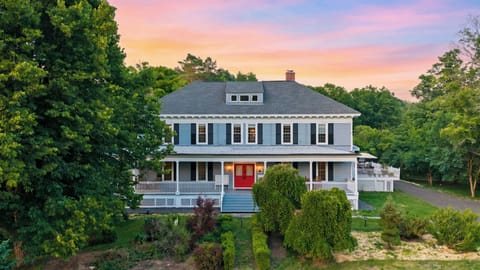 Twilight exterior showcasing the historic home’s wraparound porch, garden setting, and glowing red entry.