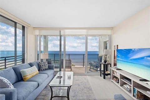 Stunning Views!  Living room with blue sofa, glass coffee table, large flat-screen TV, and sliding glass doors opening to oceanfront balcony.