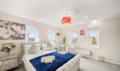 A bright bedroom features a white king bed with a blue blanket, windows, a bedside drawer, and a small workspace.