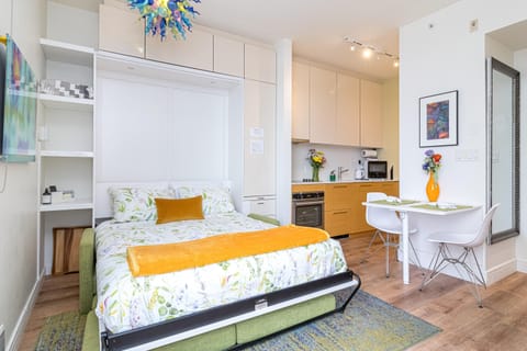 Murphy bed with floral bedding folds down over a green sofa, adjacent to white and wood kitchen cabinetry.