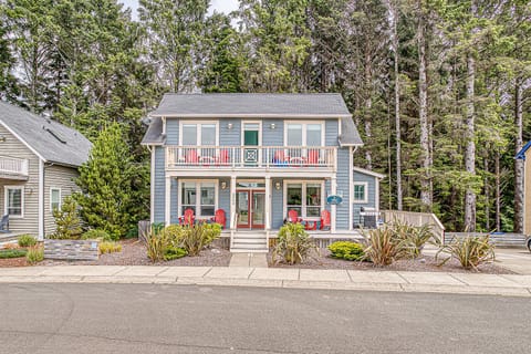 A charming blue coastal-style home nestled among tall pines, featuring bright red outdoor seating and welcoming balconies for relaxing in the fresh forest air.
