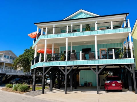 Two-story elevated beach house with pale turquoise siding, black stilts, wraparound balconies, and coral scalloped umbrellas.