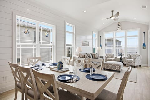 Bright open-concept great room with dining for eight, coastal living area, vaulted ceilings, shiplap walls, expansive windows, and beautiful ocean views, perfect for gathering after a day at Oak Island Beach.