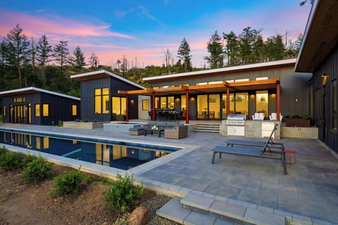 A stunning twilight shot of the resort-style backyard, featuring an illuminated lap pool, built-in hot tub, and an outdoor lounge set against a colorful evening sky.