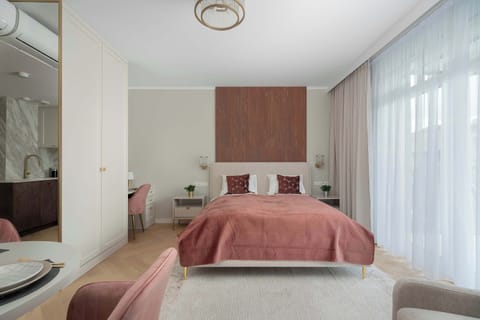 A bright bedroom features a double bed and large windows with sheer curtains. Natural light makes the space feel open and cozy.