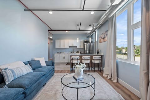 Open-concept living and kitchen with a plush blue sofa, glass coffee table, and a 3-seat bar island. White cabinetry, stainless appliances, and floor-to-ceiling windows flood the space with natural light.