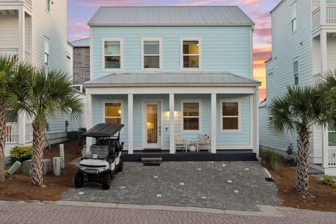 A charming coastal exterior welcomes you with a covered front porch, palm trees, and a soft sunset backdrop.