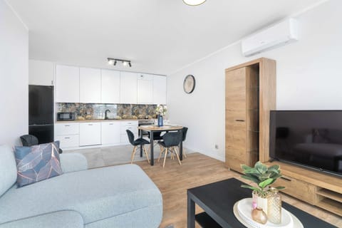 A spacious open-concept living area featuring a light grey sofa, coffee table, and open kitchen background. Bright natural light fills the room through large windows.
