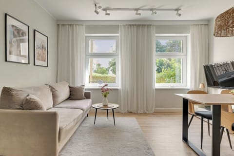 Przestronny i elegancki salon z wygodną sofą, jasną kolorystyką i dużymi oknami zapewniającymi naturalne światło. Spójna aranżacja, drewniane akcenty i przytulna strefa jadalniana tworzą komfortową przestrzeń do wypoczynku.