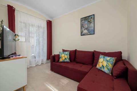 A cozy living room area with a red sofa, colorful decorative cushions, and a large window dressed with patterned curtains. A small table and framed artwork add warmth to the space.