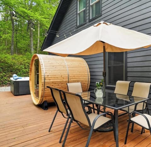 Six-seat dining table under an umbrella, barrel sauna behind it, hot tub beyond, string lights overhead, Ohio woods all around. One shot, whole story.