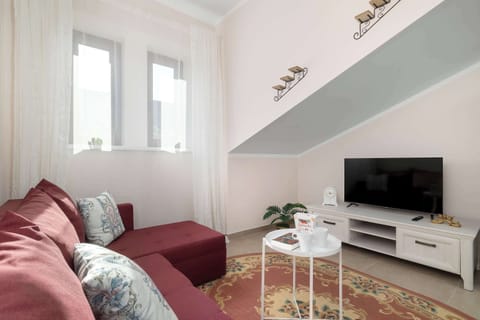A cozy living area features a dark red corner sofa adorned with cushions, positioned next to a white coffee table and a TV stand with a television. A patterned rug lies underfoot, adding warmth to the space.