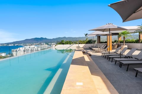 Elevated rooftop terrace featuring an expansive infinity pool, shaded sun loungers, modern umbrellas, and open mountain-to-ocean views.