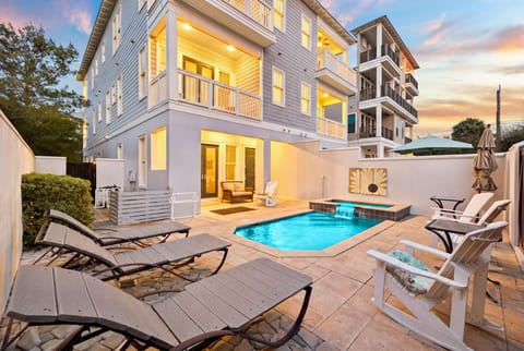 A stunning twilight view of this modern coastal property reveals your private outdoor sanctuary, complete with a sparkling pool, inviting loungers, and multiple seating areas perfect for unwinding under the evening sky.