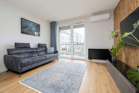 A modern living room features a dark grey plush sofa on a patterned rug opposite a wall-mounted TV framed by vertical wood slats. Sliding glass doors lead out onto a bright balcony.