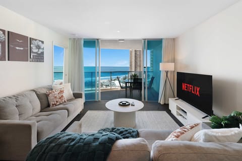 Relax in this spacious oceanfront living room, featuring plush grey sofas, a large flat-screen TV, a round center coffee table, and expansive glass doors opening to stunning beach views from the private balcony.