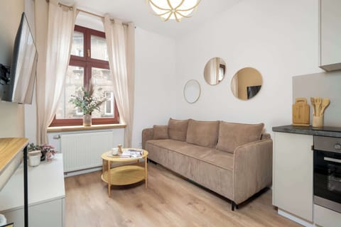 A cozy open-plan living room features a beige sofa, a round coffee table, and soft blush curtains. The kitchenette area sits to the right on light wooden floors.