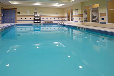 Indoor pool with clear blue water, accessible stairs, and fitness area visible behind glass.