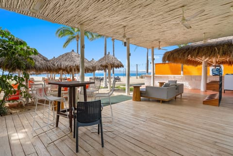 Sunlit beachfront lounge area featuring a wooden deck, casual high-top and lounge seating under a rustic thatched roof, complete with a palapa-shaded bar, open sand, and clear ocean views.