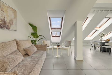 Jasny i przestronny salon na poddaszu, pełen naturalnego światła wpadającego przez okna dachowe. Komfortowa sofa, elegancka jadalnia i minimalistyczna aranżacja tworzą harmonijną przestrzeń idealną do wypoczynku i wspólnego spędzania czasu.