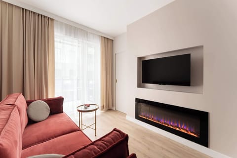 Stylowy salon z elegancką czerwoną sofą, efektownym kominkiem i telewizorem. Jasne wnętrze, naturalne światło oraz harmonijnie połączona strefa jadalniana tworzą przytulną i komfortową przestrzeń do wypoczynku.