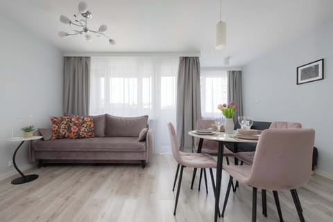 The living area features a comfortable sofa placed next to a large window with sheer curtains. On the right side, a round dining table is accompanied by elegant pastel pink chairs, creating a practical space to relax and share meals.