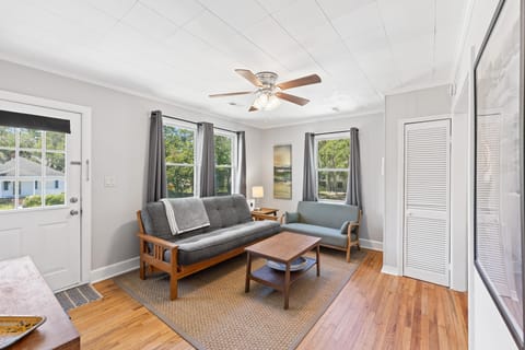 Natural light fills the living room through three windows framed by gray curtains, highlighting the hardwood floors and ceiling fan.