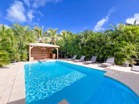 Beautiful organic-shaped pool featuring 4 grey sun loungers, natural wooden gazebo and lush garden with palm trees and tropical plants under bright blue skies