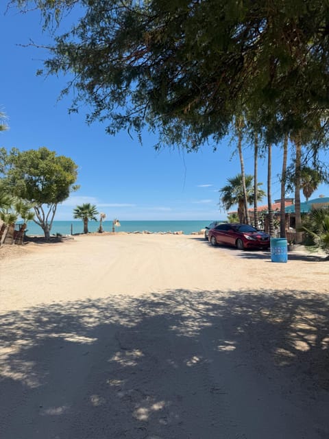 A wide outdoor view looking down the wide sandy property path toward the open blue ocean horizon, featuring parked vehicles and mature trees along the beach approach.