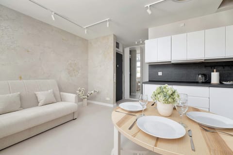 A bright open-plan living area combines a cozy sofa with a wooden dining table set for guests. Modern white cabinetry lines the kitchenette wall, creating a clean and functional living space.