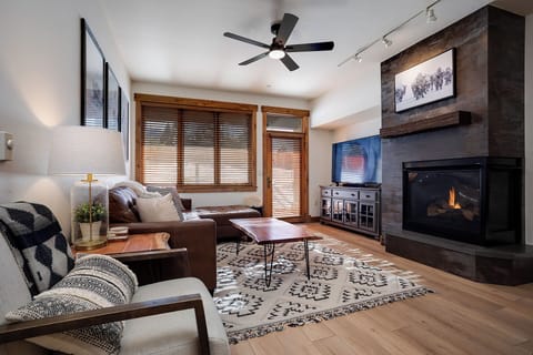 Unwind in your cozy living room, where a crackling fireplace and plush seating invite you to relax after a day of adventure. Natural light pours through wooden blinds, creating a warm mountain retreat.