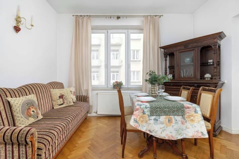 A living room features a wooden table covered with a patterned tablecloth, surrounded by matching chairs. A comfortable patterned sofa sits against the wall, next to a large window letting in natural light.