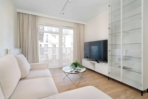 A bright living room features a beige sofa on the left and a flat-screen TV on the opposite wall. A glass coffee table sits on the light wooden floor between them.