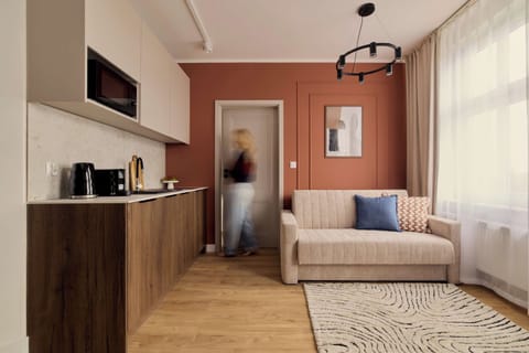 Stylowy i przytulny salon połączony z aneksem kuchennym, utrzymany w ciepłej kolorystyce. Drewniane akcenty, dekoracyjna ceglana ściana, wygodna sofa i duże okno tworzą jasną, komfortową przestrzeń do wypoczynku.