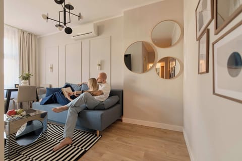Stylowy i przytulny salon z wygodną sofą, dużym telewizorem i eleganckimi dodatkami. Jasna kolorystyka, drewniana podłoga, dekoracyjne lustra oraz nastrojowe oświetlenie tworzą komfortową przestrzeń do wypoczynku i spędzania czasu.