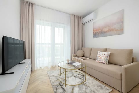 A bright living area features a comfortable beige sofa adorned with decorative pillows. A flat-screen television sits on a sleek white media console across from the seating area.