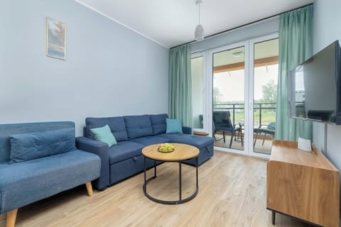 A brightly lit living room features a blue sofa, a matching armchair, and a wooden TV stand. Large glass doors lead out to a balcony, allowing plenty of natural light to enter the space.