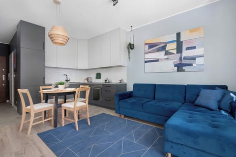 A bright living room with a blue sofa, dining table, wooden chairs, and a modern kitchen in the background. Large windows and light flooring create an open feel.