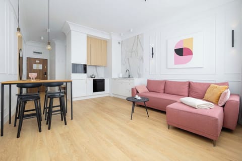 A bright open plan living area with a comfortable pink sofa and a dining table. Large windows fill the space with natural light and create a welcoming atmosphere.