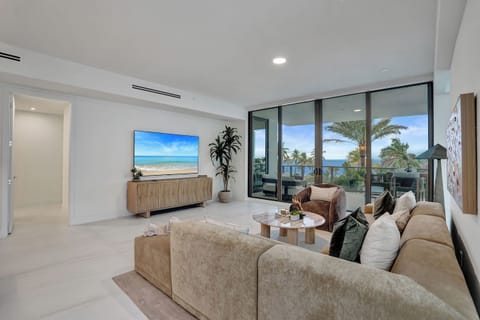 Sun-filled living room with sectional sofa, large TV, and floor-to-ceiling glass framing palm trees and ocean.