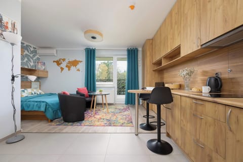 A bright studio with a comfortable double bed, a kitchenette, and a small dining area. Large windows and blue curtains bring plenty of natural light into the space.