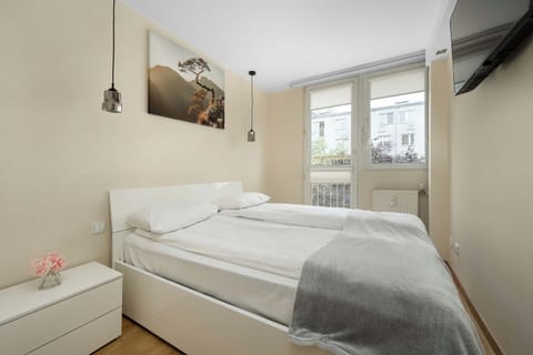 A bright, compact bedroom with a neatly made double bed covered in white and light-gray bedding. A large window brings in natural light, while a wall-mounted television and a small hanging light fixture are visible.
