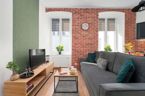 A bright living room with a gray sofa, a small coffee table, and a dining area near the windows. Exposed red brick walls, large windows, and green accents create a warm, modern atmosphere.