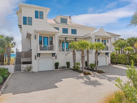 Modern beachfront villa with elegant coastal architecture and tropical palm landscaping, featuring a private paved driveway and garage access.